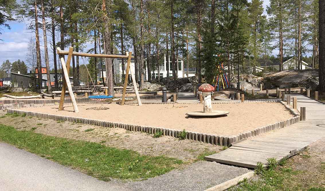 Överbilck av lekplatsen. En komisgunga och lekgunga med sandunderlag. Träramper leder runt lekplatsen. I bakgrunden synt upplagda stockar i olika höjder och klätterställnig.