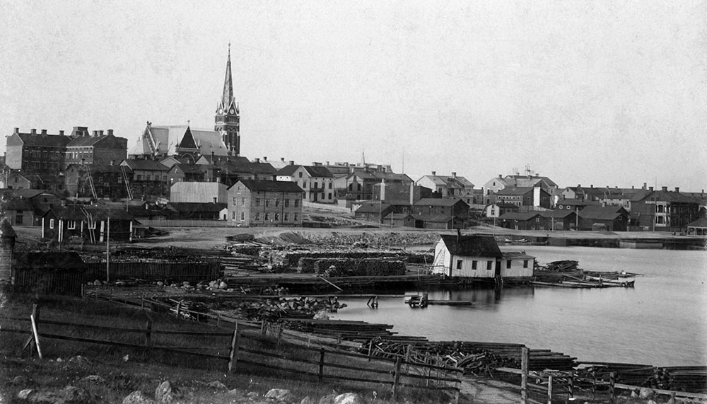 Vy över hamn och stad med litet hus och brygga i förgrunden.