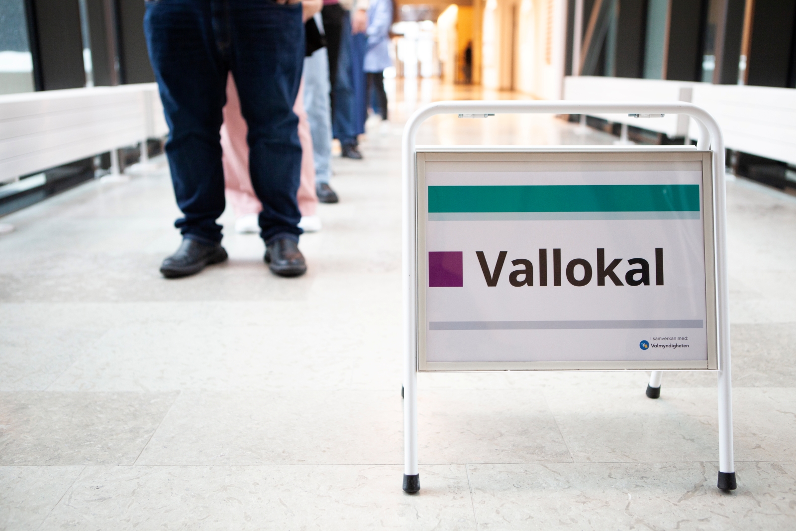 Skylt med texten "vallokal". I bakgrunden står personer i ran för att rösta.
