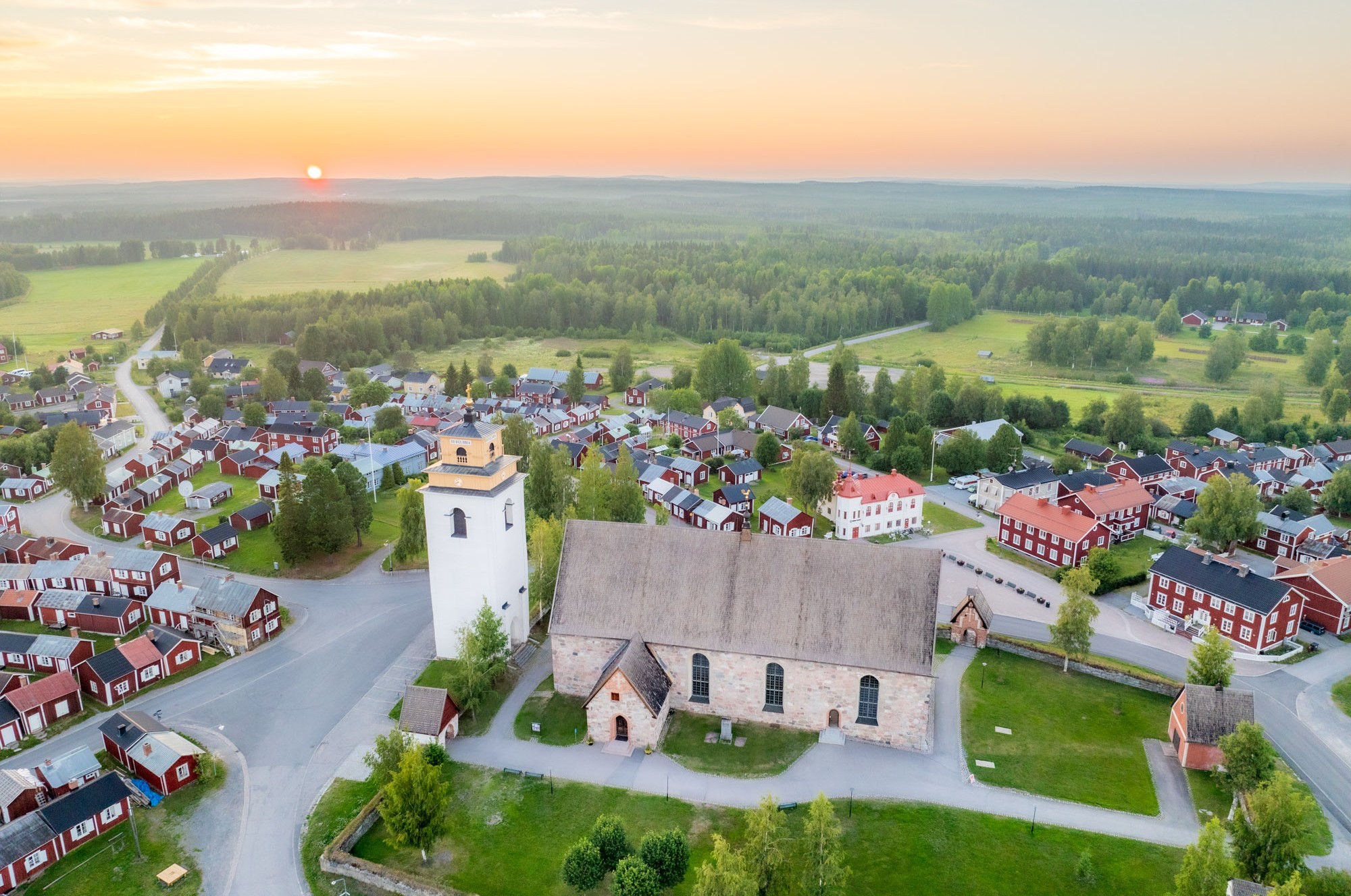Världsarvet Gammelstads kyrkstad från luften. Kyrka med små stugor och vägar runtomkring. Gul himmel.