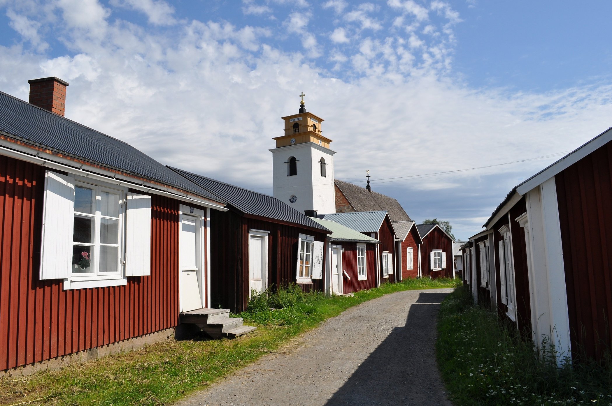 Väg med små röda och vita kyrkstugor i Gammelstads kyrkstad. Vitt kyrktorn i bakgrunden. Blå himmel och vita moln.
