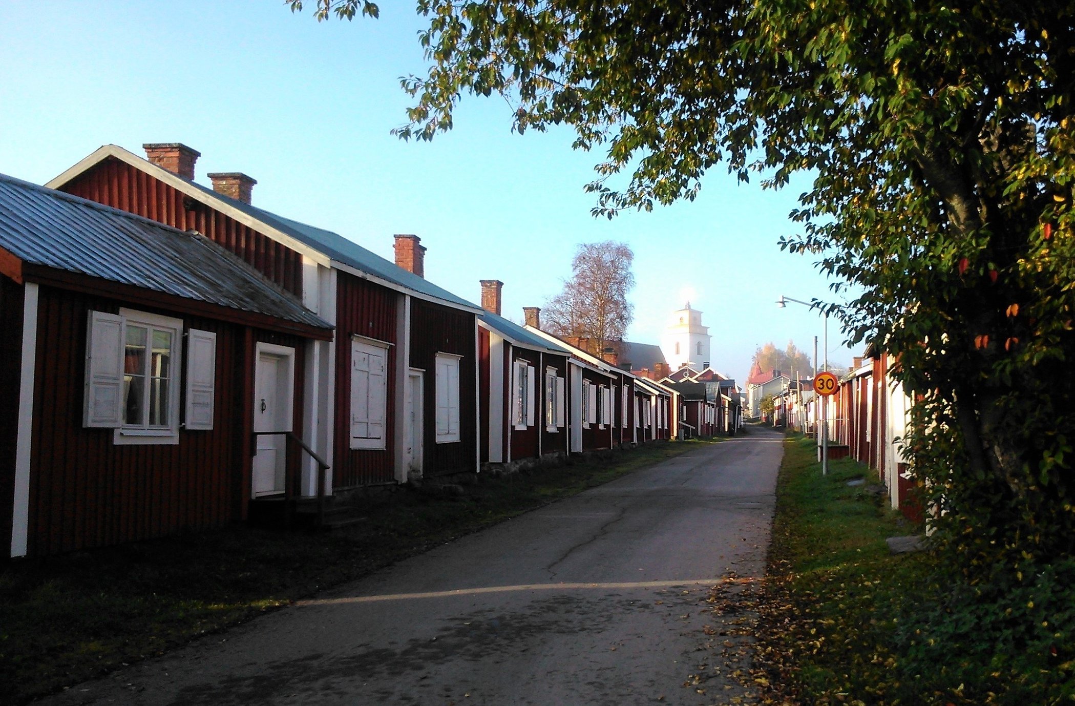 Väg med små röda och vita kyrkstugor i Gammelstads kyrkstad. Trädgrenar i förgrunden.
